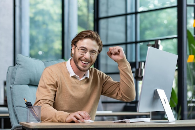 幸せな若い男子学生がオフィスの自宅のコンピューターの前に座って、勝利を示しています