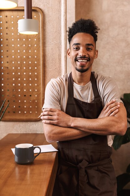 Felice giovane barista che indossa un grembiule in piedi presso la caffetteria, con le braccia conserte