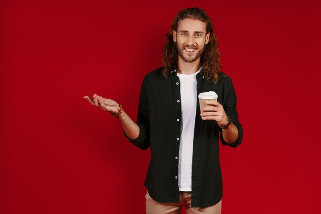 Happy young man in casual clothing looking at camera with smile and pointing copy space while standing against red wall