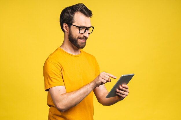 Happy young man in casual standing and using tablet computer isolated over yellow background.