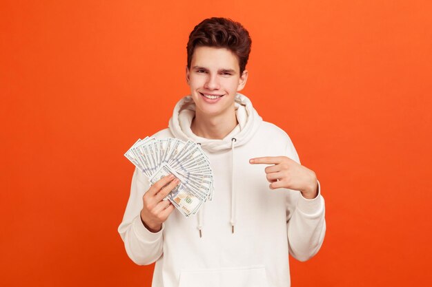 Happy young man in casual style hoodie pointing finger on bunch of dollars cash with toothy smile on face, good salary on perspective job. Indoor studio shot isolated on orange background