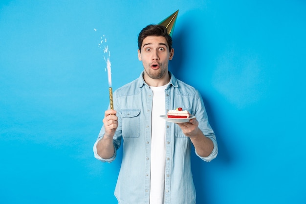 Счастливый молодой человек празднует день рождения в партийной шляпе, держит торт на день рождения и улыбается, стоя на синем фоне
