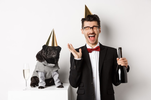 Felice giovane che celebra la vacanza con il simpatico cane, con in mano champagne e sorridente, carlino e proprietario che indossano costumi da festa, bianchi.