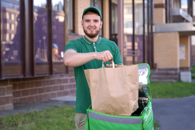 Фото Счастливый молодой курьер в зеленой форме с большой термо сумкой или рюкзаком доставляет еду из