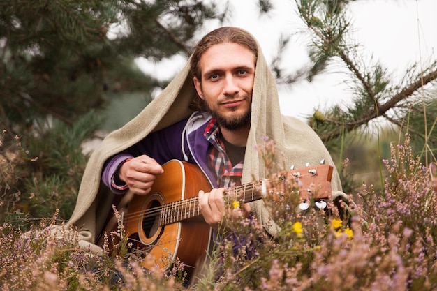 Happy young man covered by plaid playing acoustic guitar and sing song. Man in pine tree forest with heather flowers meadow. Close up, copy space.