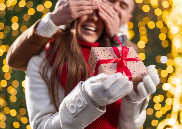 Foto giovane felice che copre gli occhi della fidanzata amata eccitata mentre fa sorpresa e dà il regalo di natale vicino all'albero incandescente