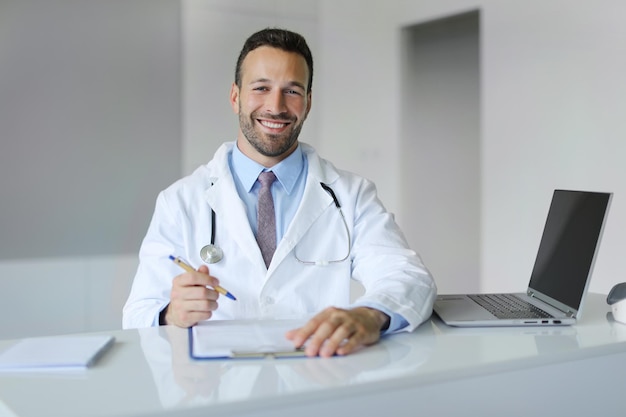 Happy young man doctor in medical coat working at his office at modern clinic writing prescription