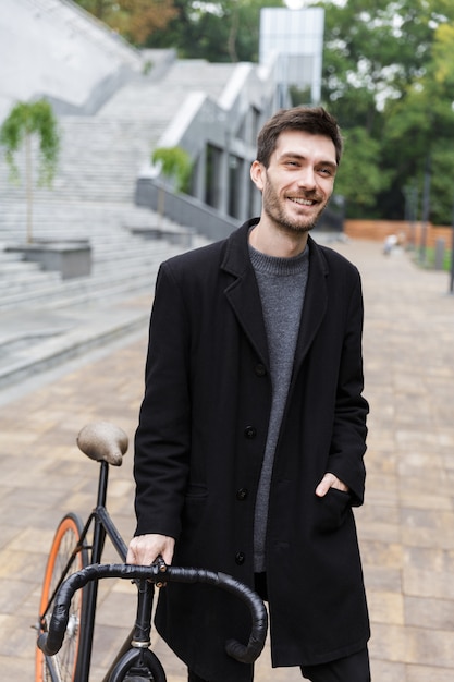 Happy young man dressed in coat walking with a bicycle on a street