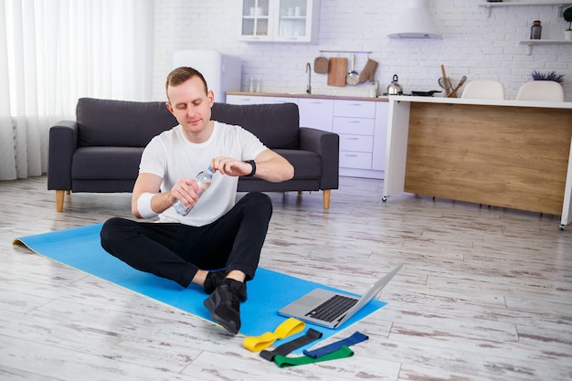 Happy young man drinks water after training online. Doing sports at home, healthy lifestyle