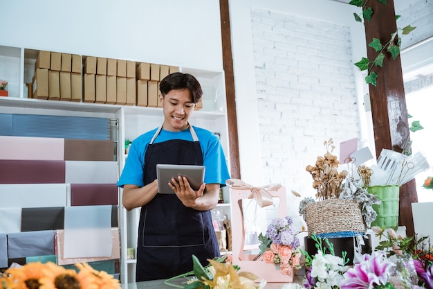 Happy young man entrepreneur working in flower shop wearing apron smiling while holding tablet