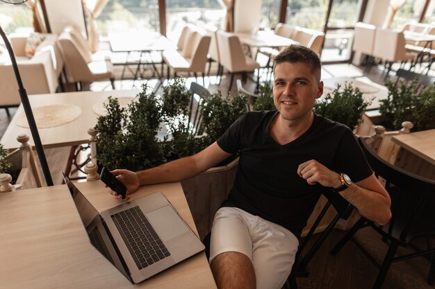 Happy young man in a fashionable black t-shirt is resting while sitting at a table in a vintage cafe with a modern laptop and with a phone in hands. Successful joyful freelancer guy relaxes indoors.