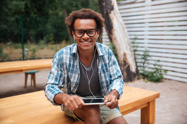 Happy young man in glasses and earphones using tablet in park