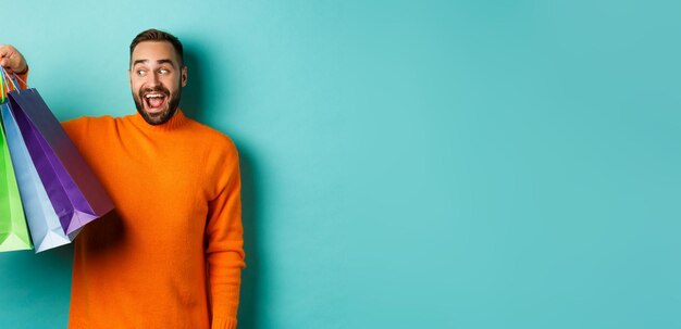 Happy young man going shopping holding bags and looking excited standing in orange sweater over person