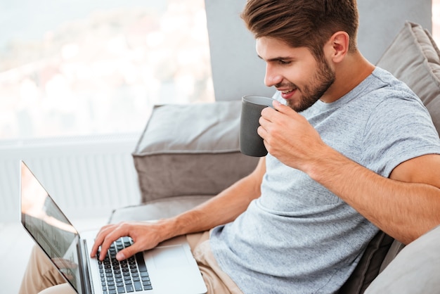 Happy young man in grey t-shirt sitting on sofa at home. Working on laptop computer and smiling while drinking a coffee.