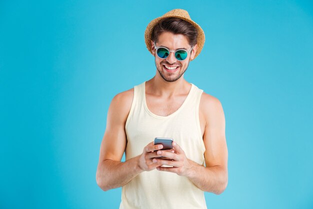 Happy young man in hat and sunglasses using smartphone over blue wall