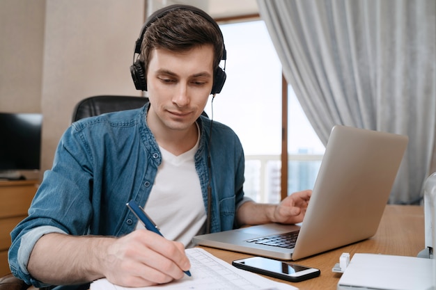 Happy young man in headphones sitting at desk in home office, writting on paper