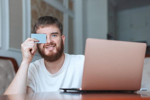 幸せな若い男がクレジット バンク カードを手に持って、自宅でラップトップ コンピューターを使用して笑顔でオンライン ショッピングをする