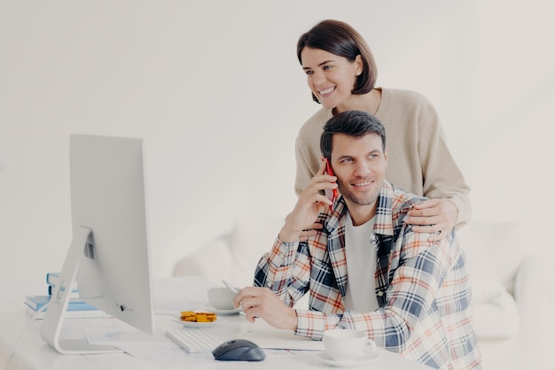 Happy young man holds mobile phone discusses issues manages\
home accounts with wife pose in coworking space sit in front of\
computer discuss family expenses couple collaborate together