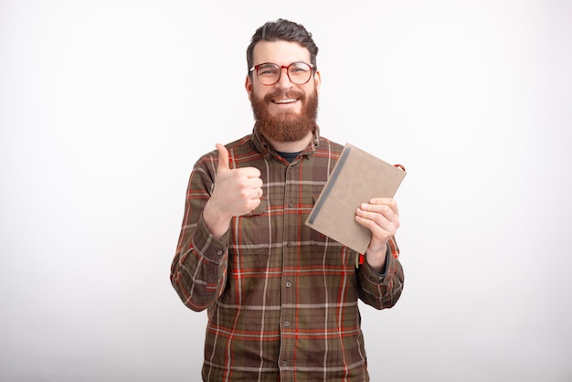 Il giovane felice sta tenendo un taccuino o un libro, sta sorridendo alla macchina fotografica, sta mostrando il bottone o il pollice simile su su spazio bianco.