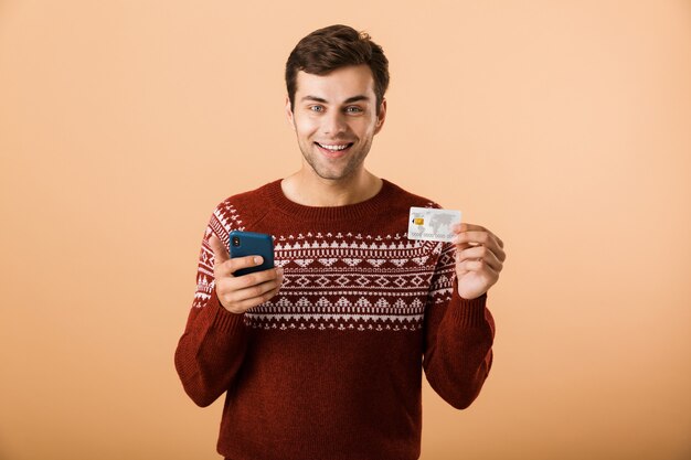 Happy young man isolated over beige wall using phone holding credit card