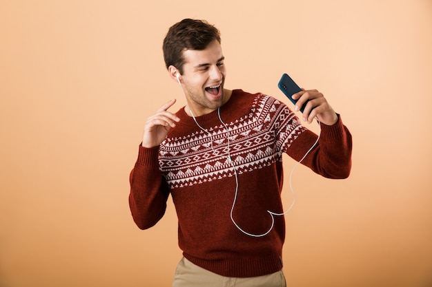 Happy young man isolated over beige wall using phone, listening to music by earphones and singing