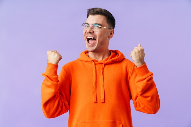 happy young man isolated over purple wall wearing eyeglasses showing winner gesture.