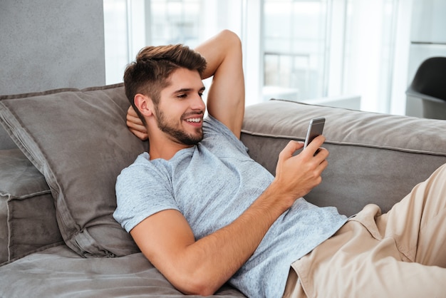 Felice giovane uomo si trova sul divano e alla ricerca sul telefono