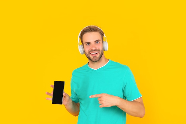 Счастливый молодой человек слушает музыку в наушниках, указывая пальцем на мобильный телефон на желтом фоне смартфона