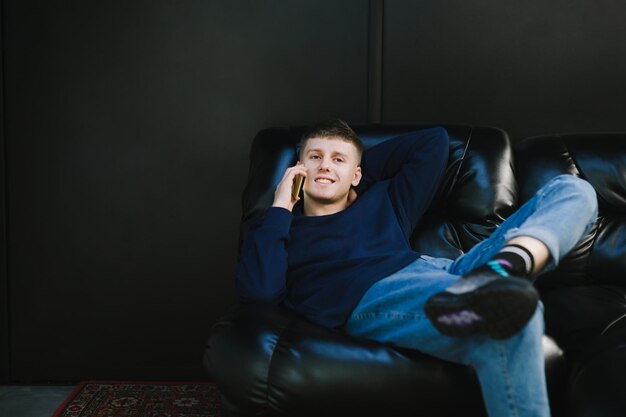 Happy young man lying on black couch against black wall background talking on phone