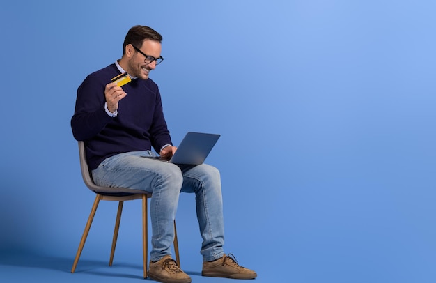 Счастливый молодой человек делает онлайн-платеж за услугу с ноутбуком и кредитной картой на синем фоне