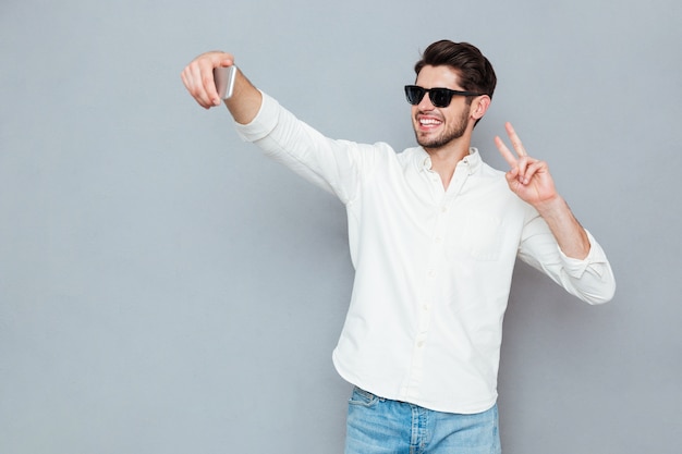 Happy young man making selfie photo on smartphone over gray background