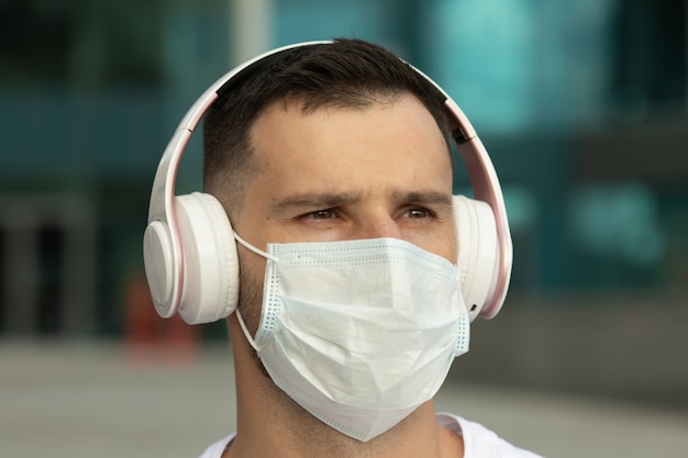 保護医療フェイスマスクの幸せな若い男は、ワイヤレスブルートゥースイヤホンで音楽を聴きます。 COVID-19コロナウイルス。