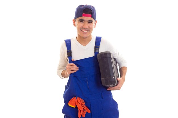 Happy young man in shirt and blue overall with orange gloves and blue snapback holding toolbox