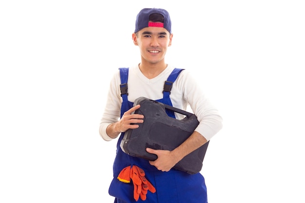 Happy young man in shirt and blue overall with orange gloves and blue snapback holding toolbox