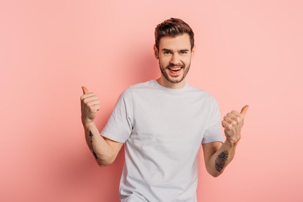 Happy young man showing thumbs up while smiling at camera on pink background