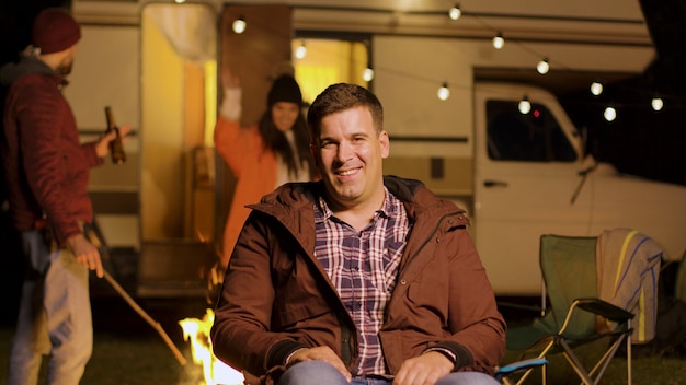 Happy young man sitting on camping chair looking at the camera in a cold night of autumn. Retro camper.