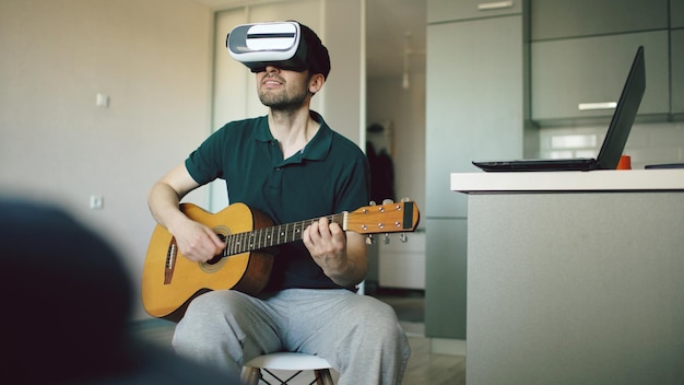 VR 360 헤드셋을 사용하여 기타 연주를 배우는 부엌에 앉아 있는 행복한 청년