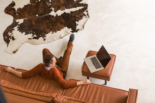 Happy young man sitting on sofa at home, working on laptop computer, smiling.