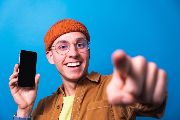 Photo happy young man smiling showing smartphone with mobile app clean screen for mockup pointing at you