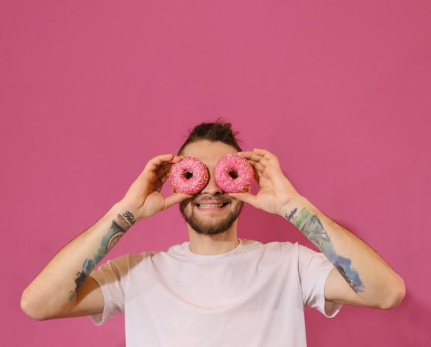 그의 눈 앞에서 두 개의 분홍색 도넛을 들고 재미 동안 웃는 행복 한 젊은 남자