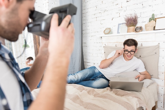 Happy young man taking photo of his handsome boyfriend posing and lying in bed with laptop.