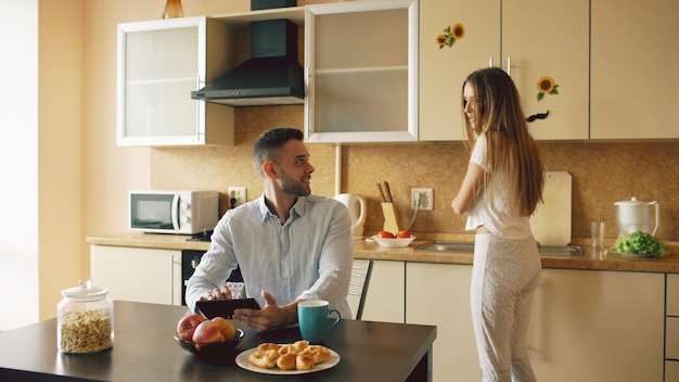 Счастливый молодой человек с цифровым планшетным компьютером сидит на кухне и разговаривает со своей женой