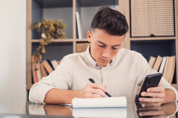 집에서 스마트폰을 사용하는 행복한 청년은 실내에서 혼자 즐겁게 숙제를 하며 인터넷 서핑을 합니다. 전화를 가진 남자 십대는 공부하고 일하는 자유 시간을 즐깁니다.