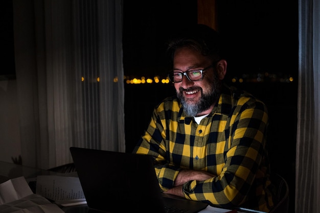 Giovane felice che usa un laptop o un computer a casa che lavora a tarda notte al buio con la luce dello schermo sul viso sorridentexa