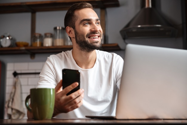 Happy young man using laptop computer while sitting at the kitchen table, holding mobile phone