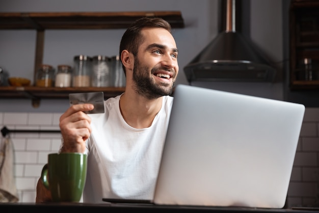 Happy young man using laptop computer while sitting at the kitchen table, showing credit card