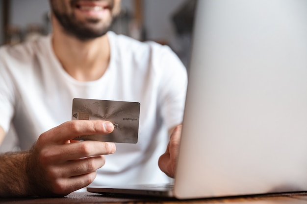 Happy young man using laptop computer while sitting at the kitchen table, showing credit card