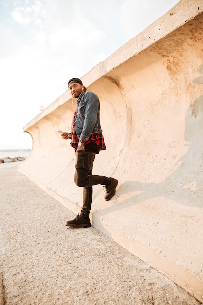 Happy young man walking on beach and chatting by his phone while listening music with earphones. Looking aside.