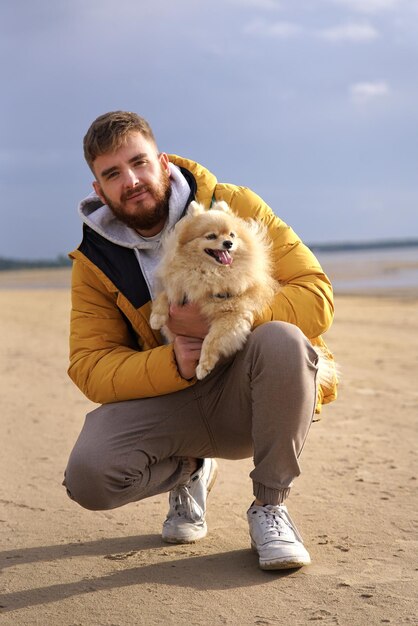 Photo happy young man walking with his pet pomeranian spitz dog on the beach hold puppy on hands people