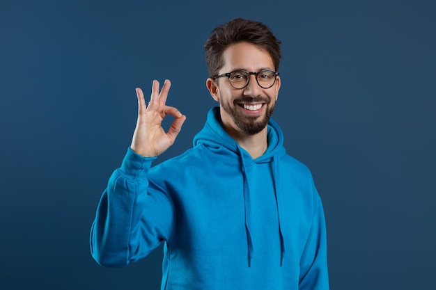 Happy young man wearing blue hoodie showing ok gesture at camera